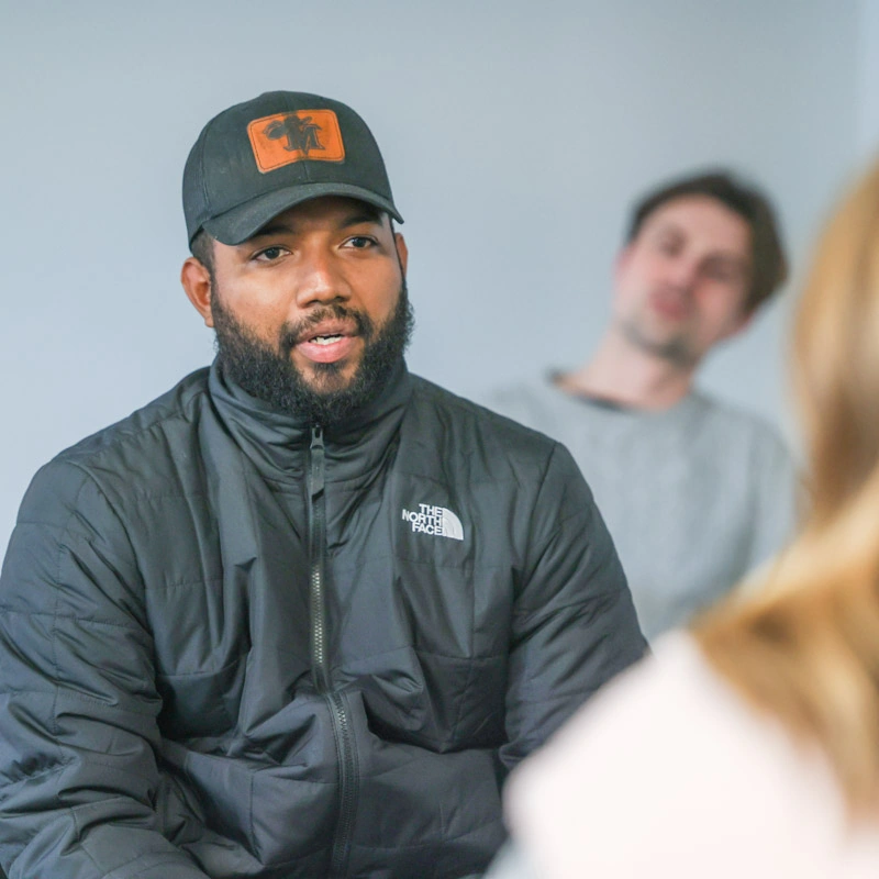 A man in a baseball cap and jacket faces a woman who is out of focus in the foreground