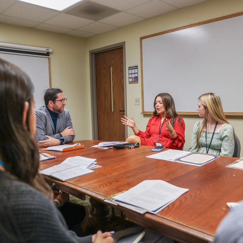 Men and women sit around the conference table in the Belk conference center as a woman wearing a bright red jacket speaks and gestures with her hands